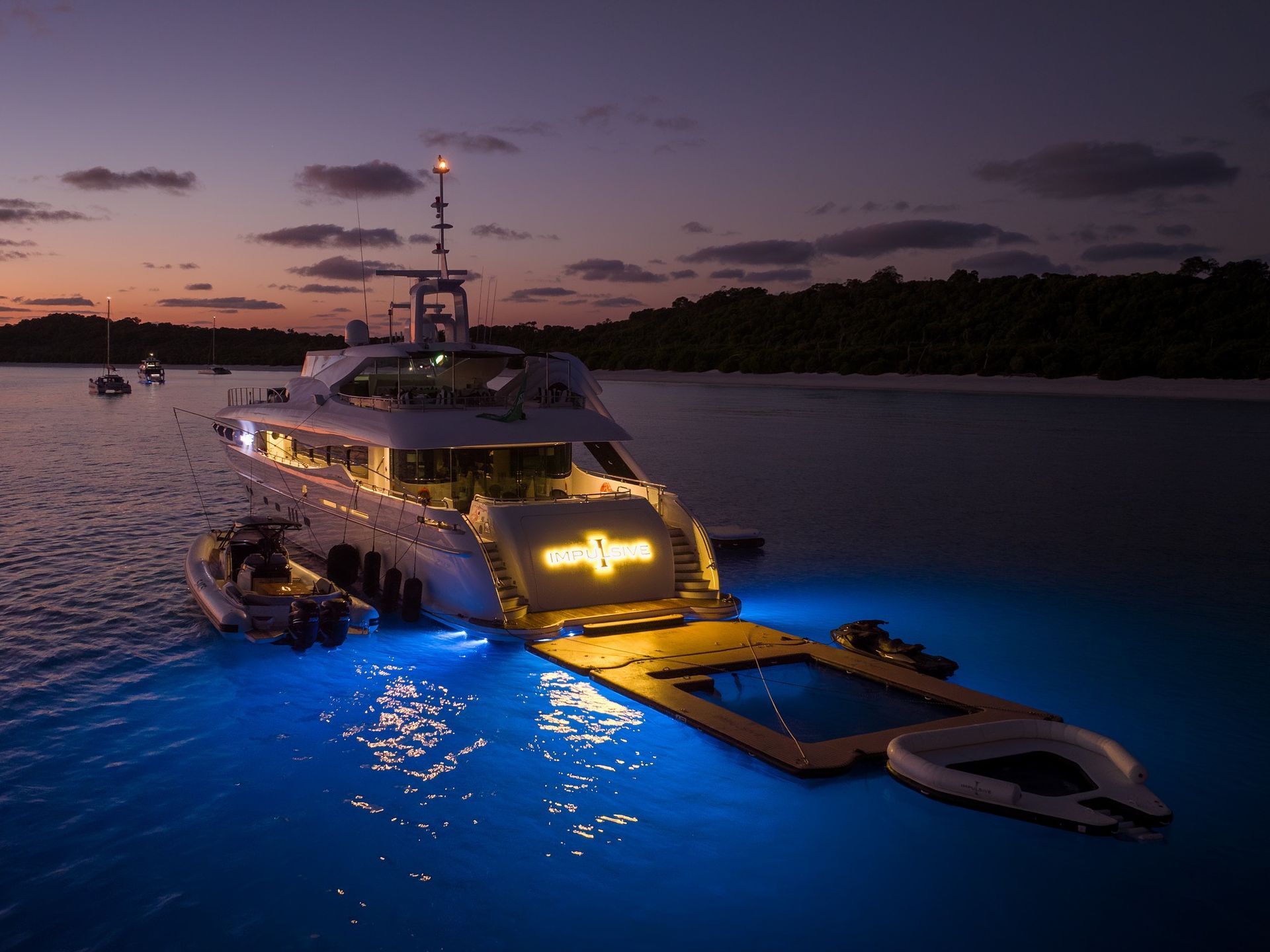 A large, illuminated yacht anchored in a calm sea at twilight, with blue underwater lights and an inflatable pool deck.