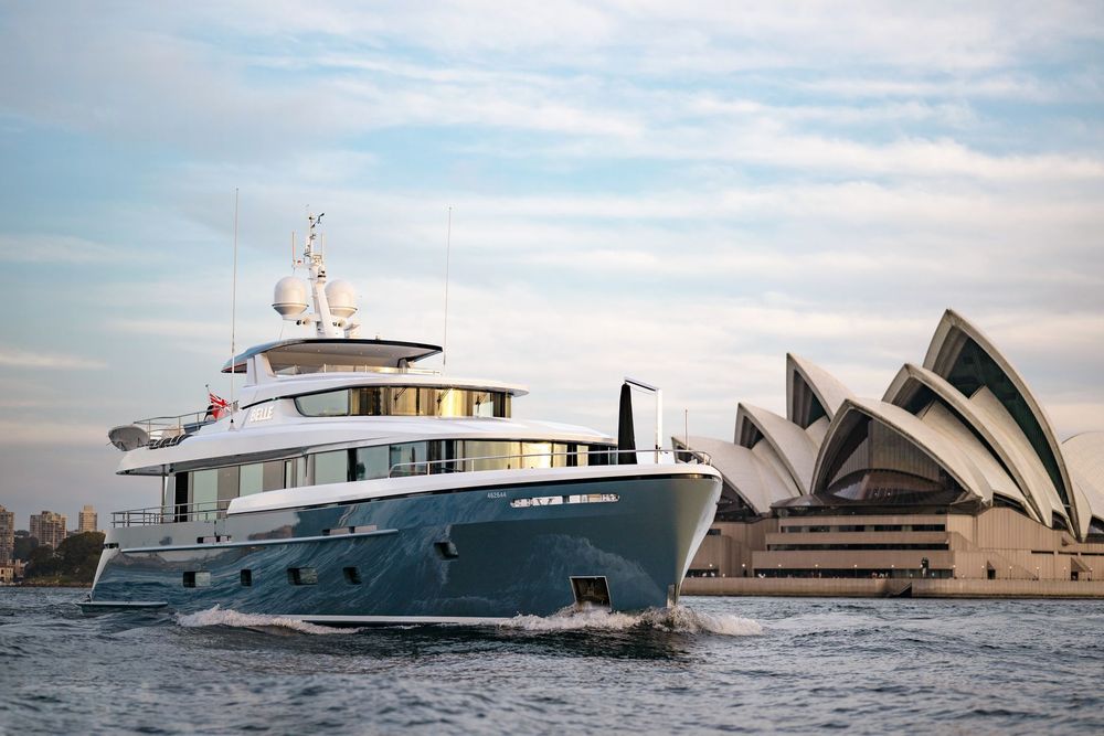 A modern yacht with a dark hull sails past the Sydney Opera House during the day.