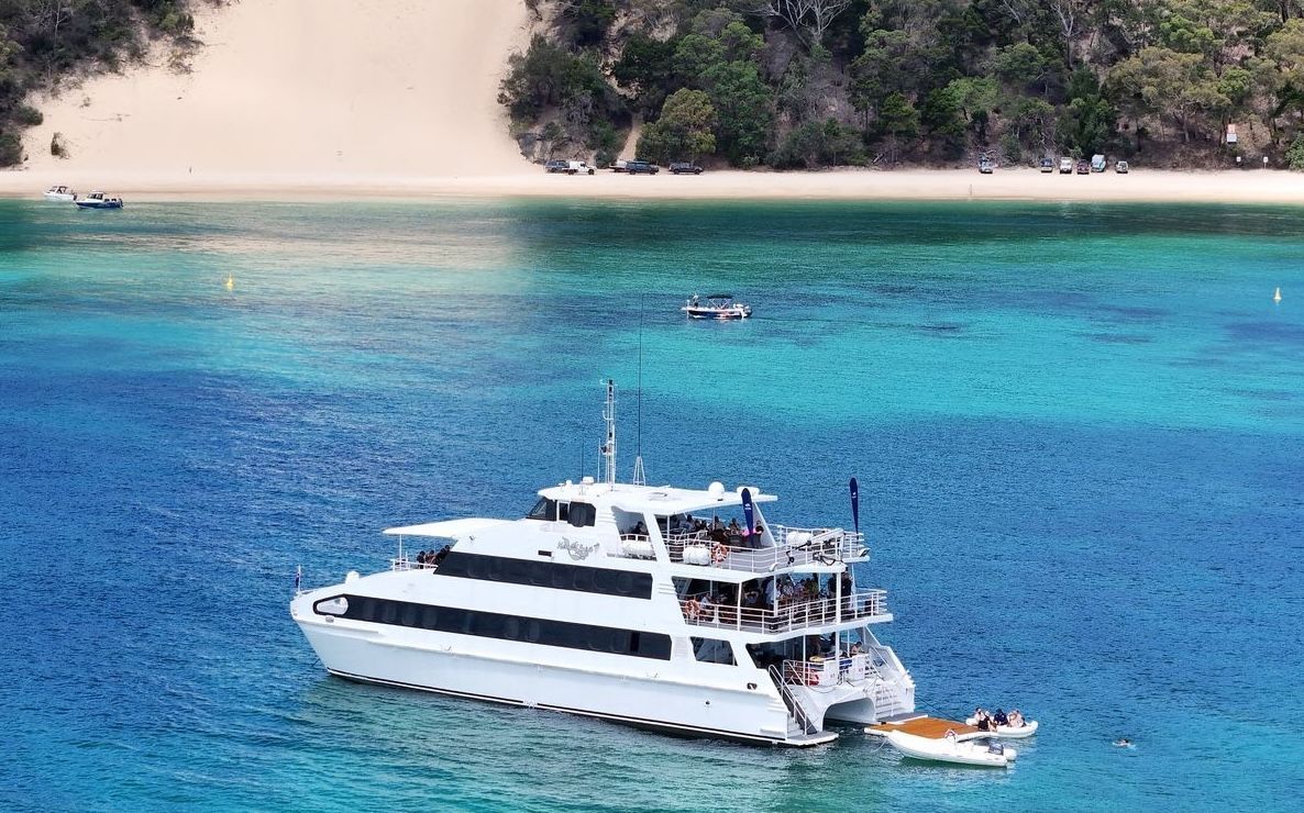 A large white catamaran cruise boat anchored in clear turquoise water near a sandy beach and green hills.
