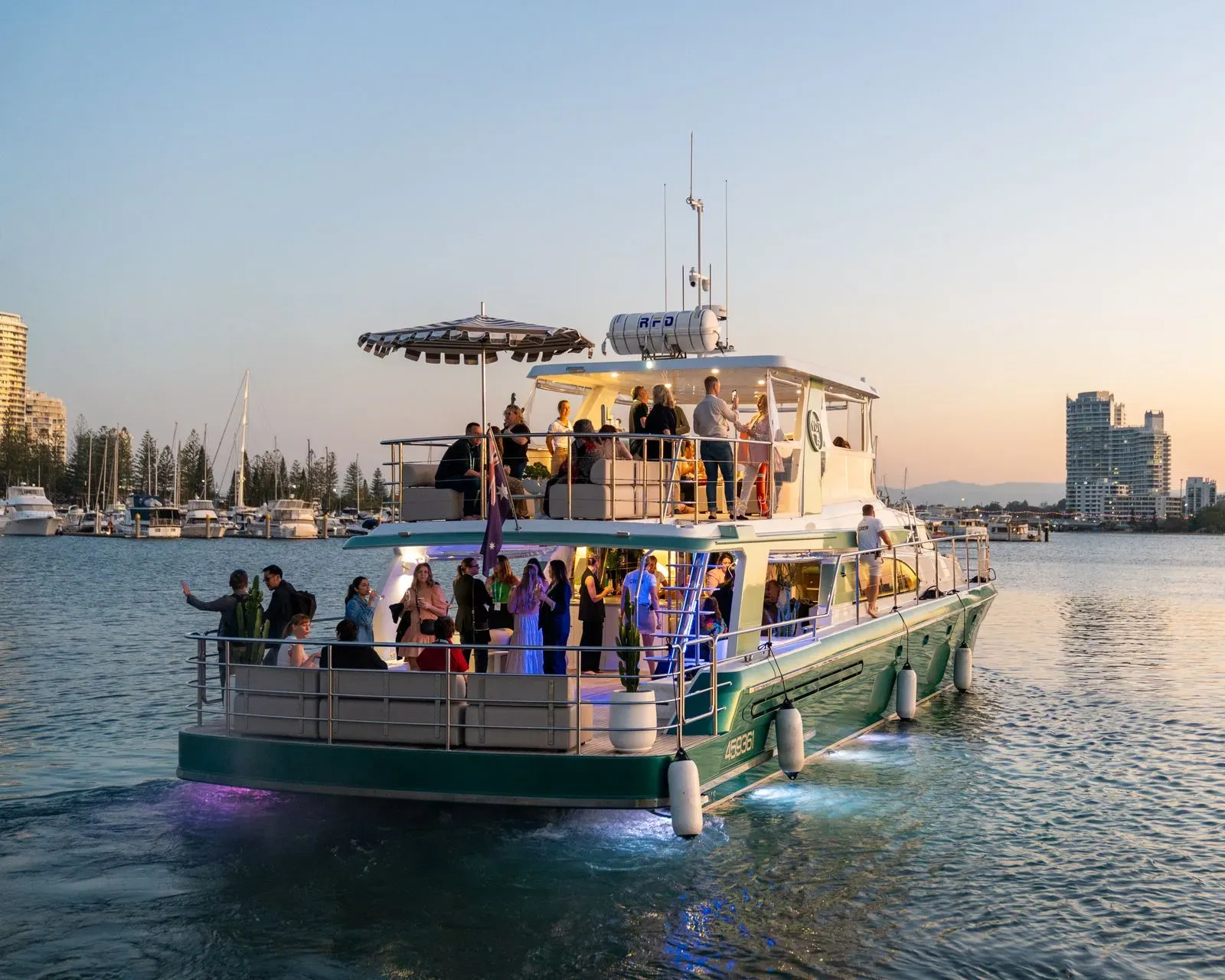 A large, multi-level cruise boat carries people on an evening excursion in a harbor near city buildings at sunset.