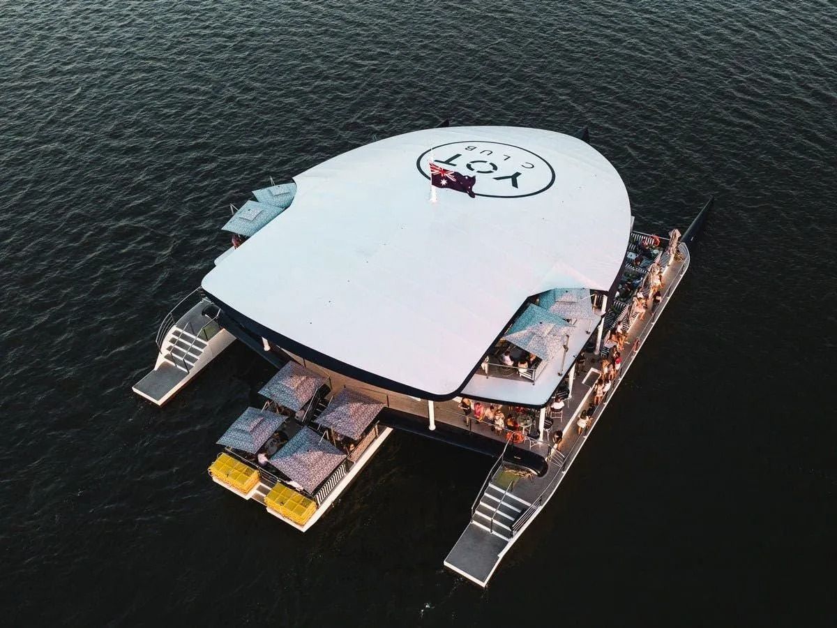 Aerial view of a white multi-level party catamaran boat floating on dark water with a large circular logo on its roof.
