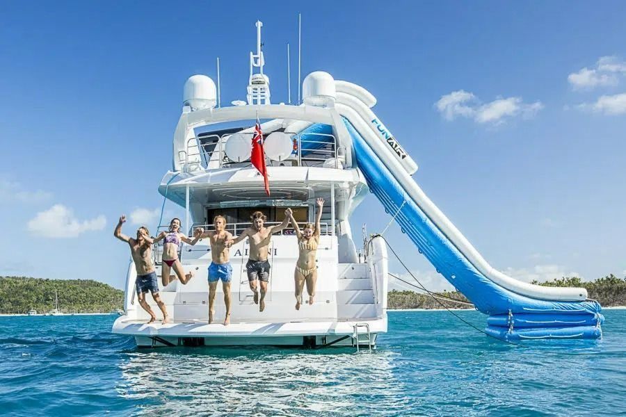 Five people jump off the back of a white yacht into turquoise water next to a large blue and white inflatable slide.
