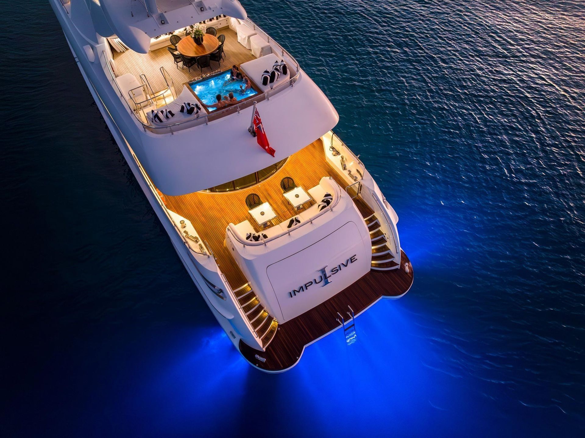 Aerial view of a luxury yacht at night, with illuminated decks, a small pool, and blue underwater lights in the sea.