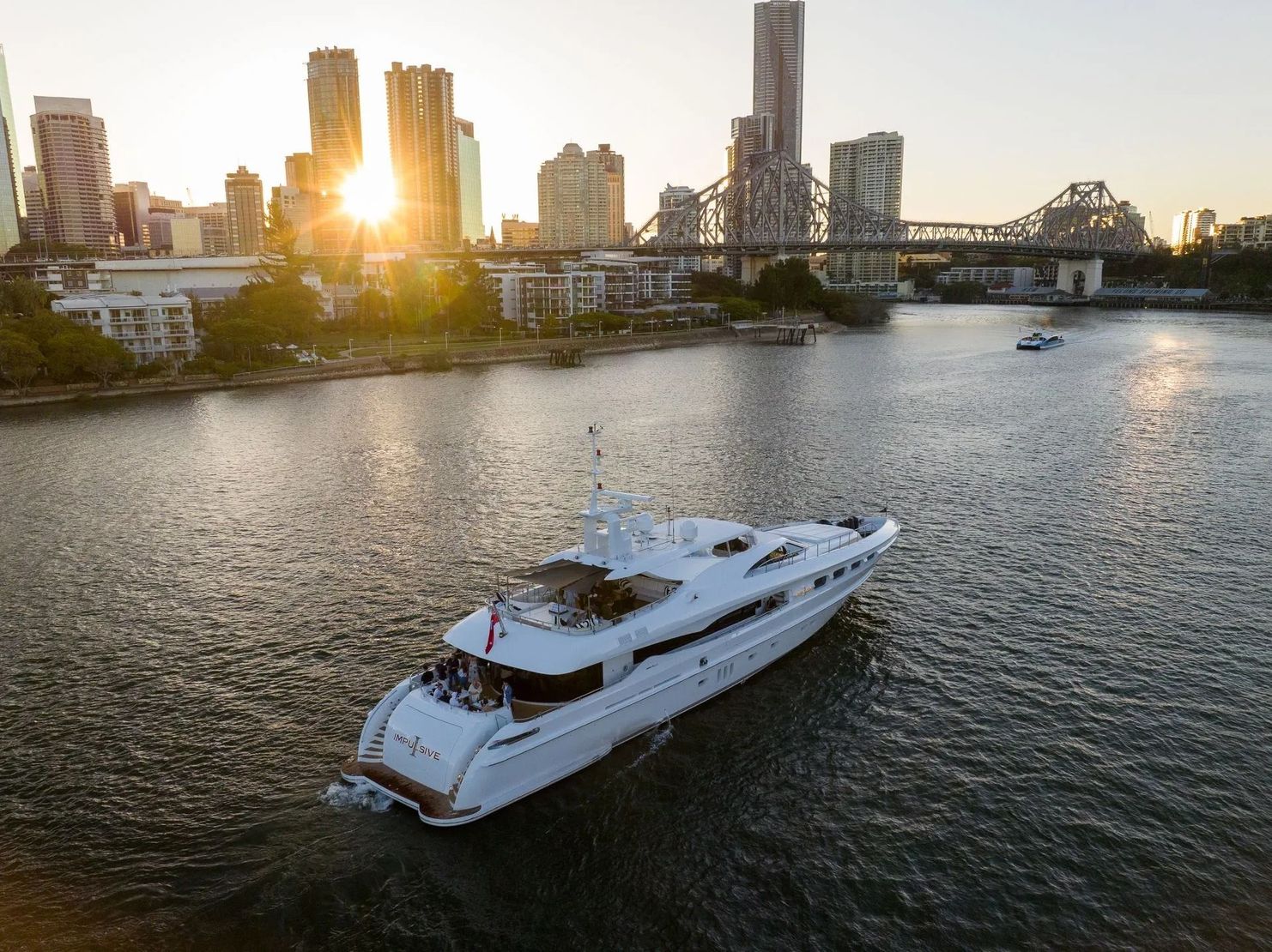 A group of people socialize on a green-and-white multi-level boat cruising on calm water near a city at sunset.