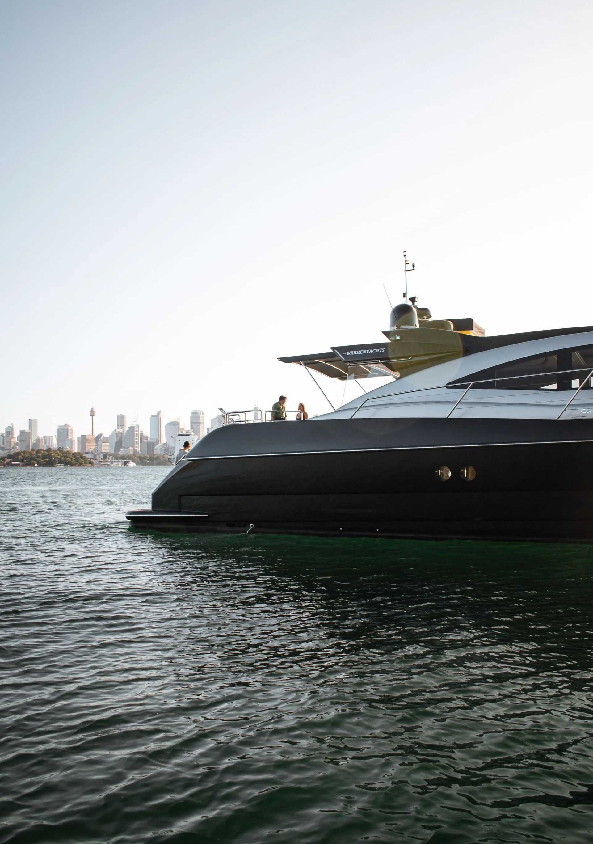 A black luxury yacht moored in a harbor with a cityscape visible in the distance under a bright sky.