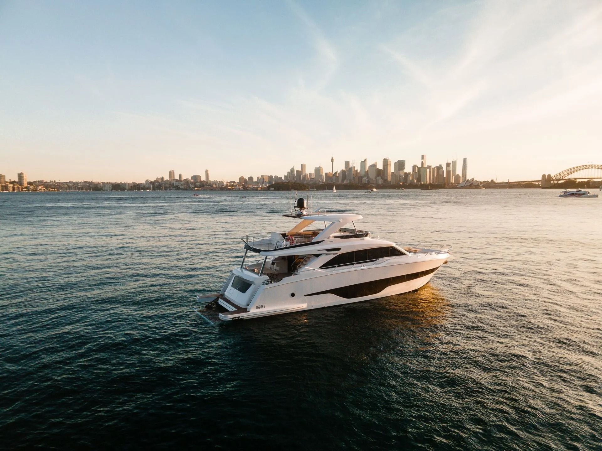 A sleek white motor yacht cruises on calm water with the Sydney skyline and Harbour Bridge in the background at sunset.