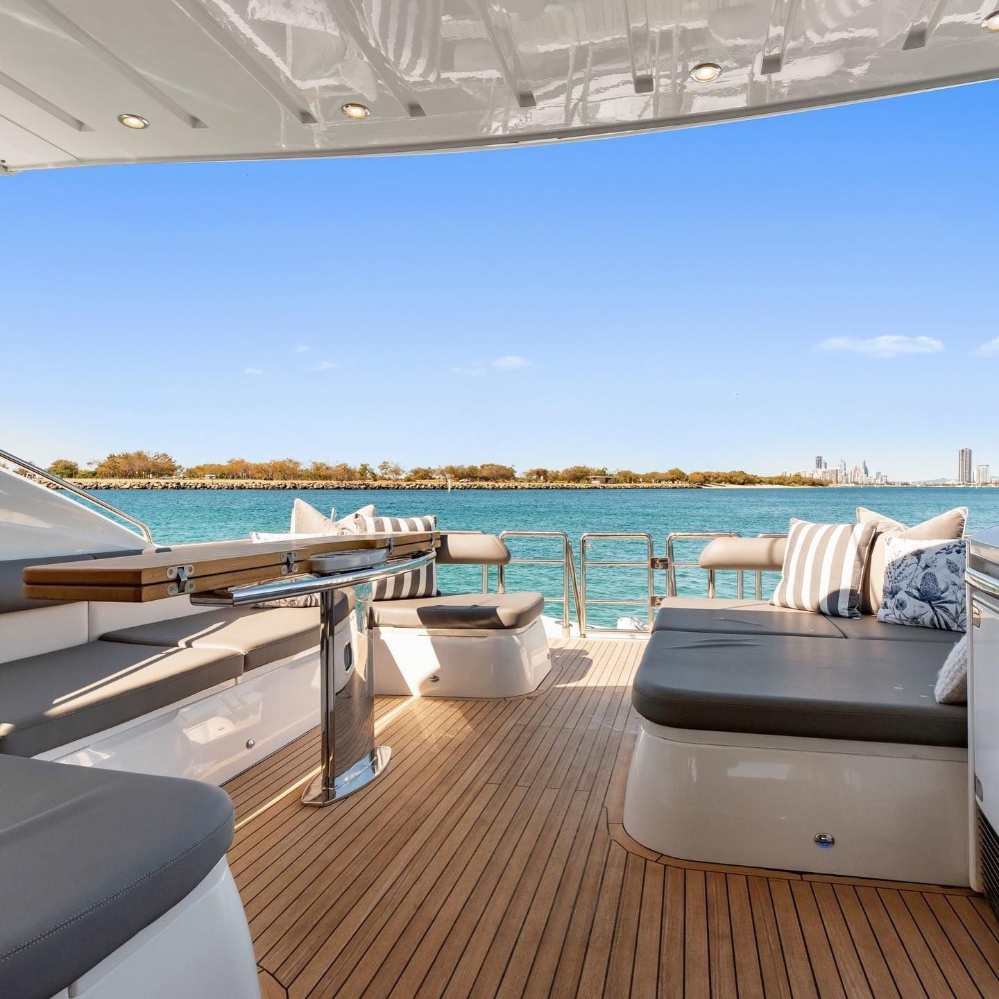 Sunlit yacht deck with wooden floors, grey cushioned seating, and open water views under a clear blue sky.