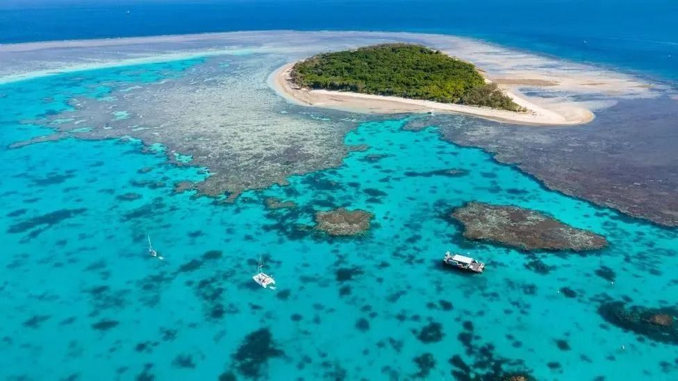 An aerial view of a green vegetated island surrounded by a coral reef and bright blue ocean waters with two boats.