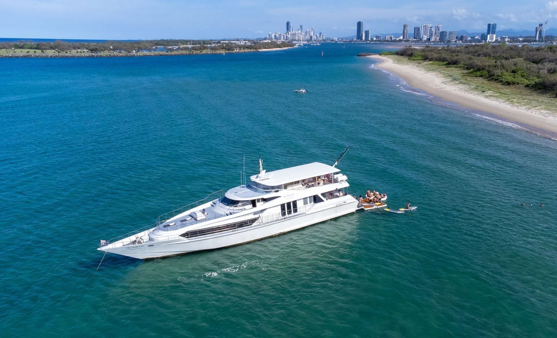 Aerial view of a white yacht anchored in clear blue ocean water near a sandy beach with a city skyline in the distance.