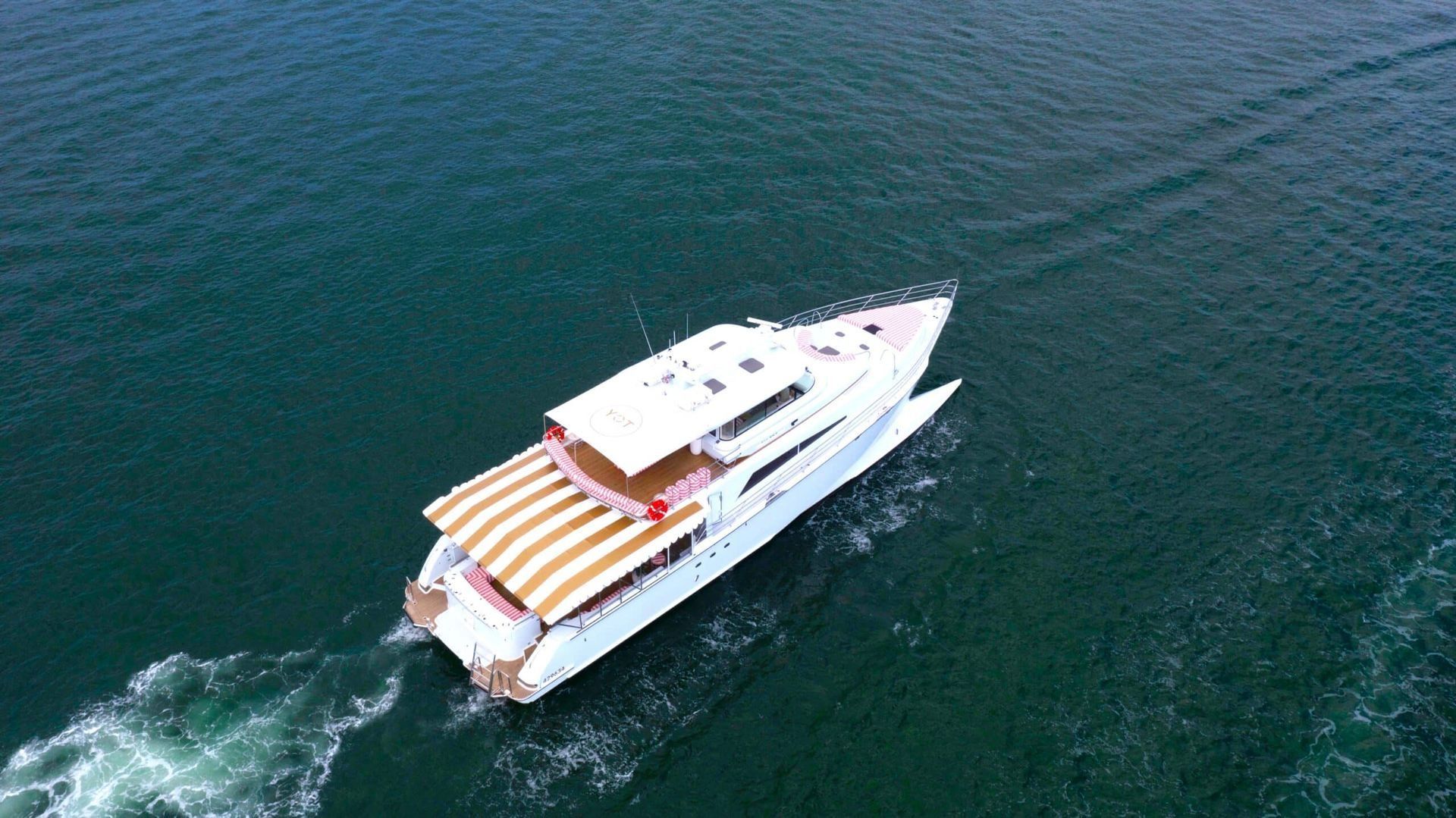 An aerial view of a white yacht cruising through dark blue water, featuring a striped, tan and white aft deck awning.