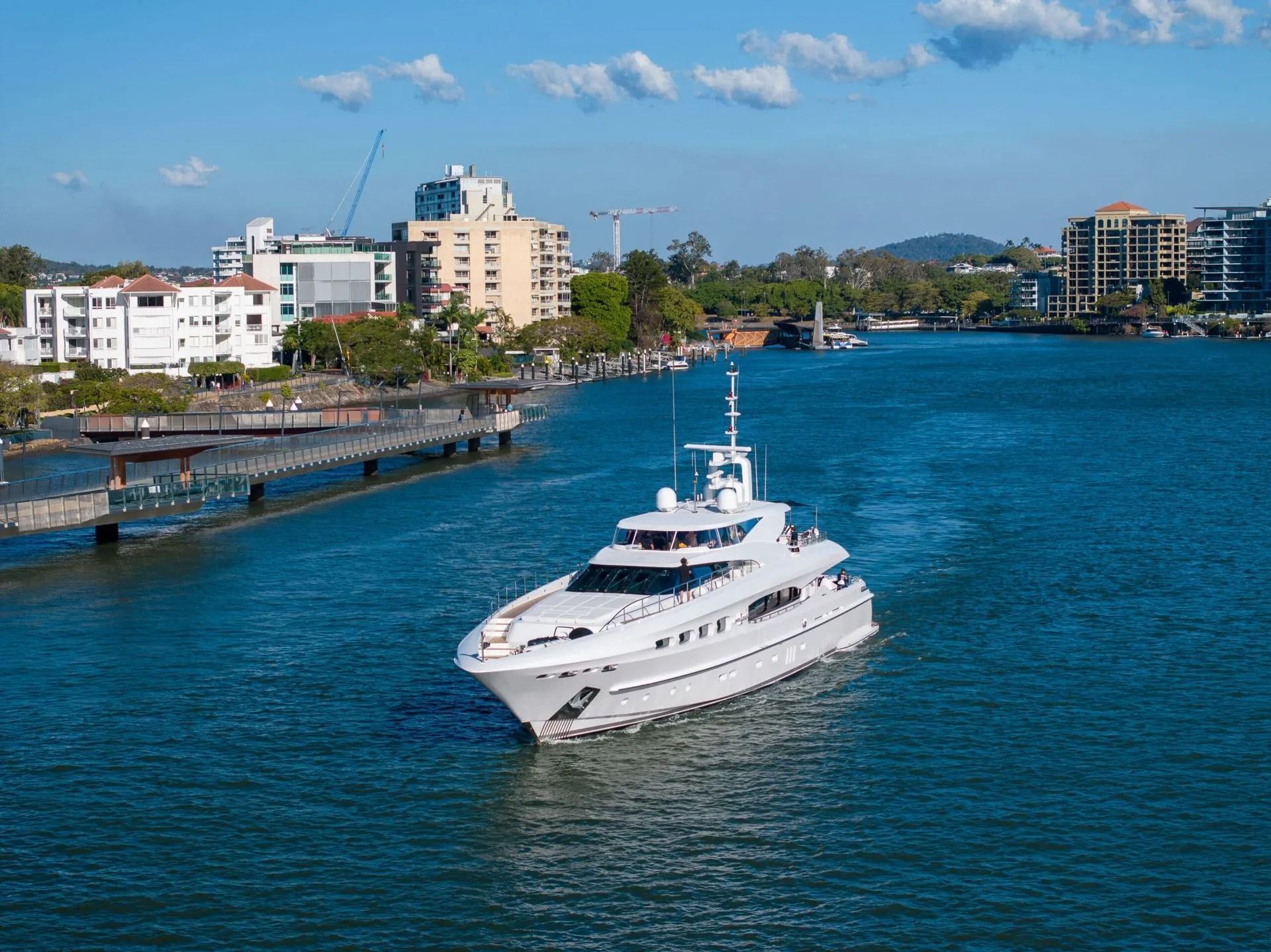 A large white yacht cruises down a sunny river flanked by modern apartment buildings and green trees.