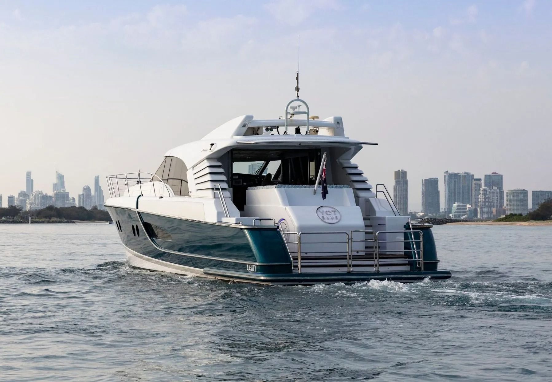 A sleek, dark teal and white motor yacht cruising on calm water, with a blurred city skyline in the background.