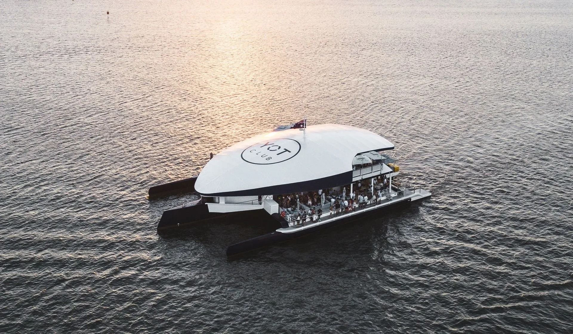 A white, futuristic, oval-shaped boat with a deck of people sits on the calm, sparkling surface of a large body of water.