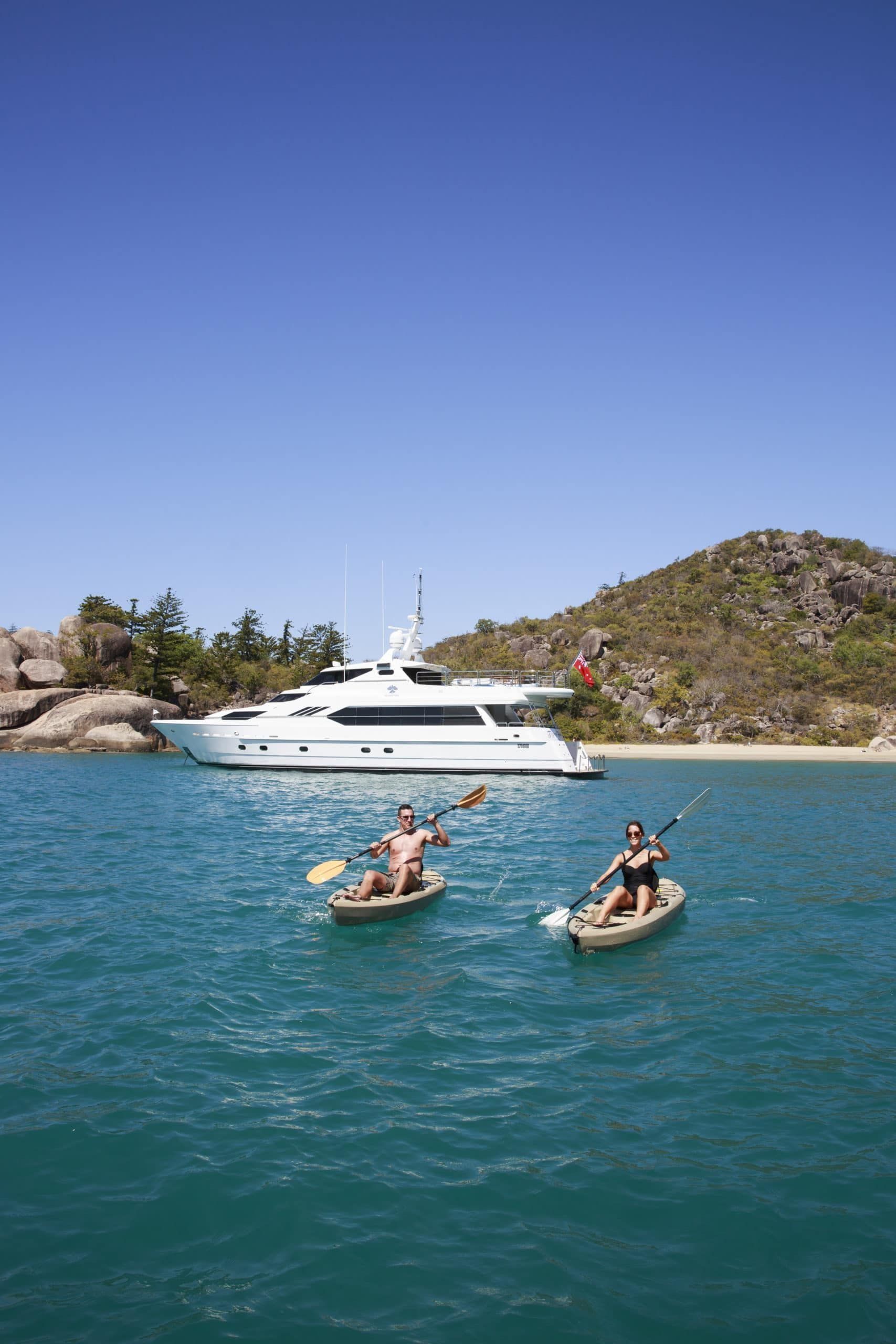 Two people in kayaks paddling on clear blue water with a large white yacht anchored in front of a coastal island.