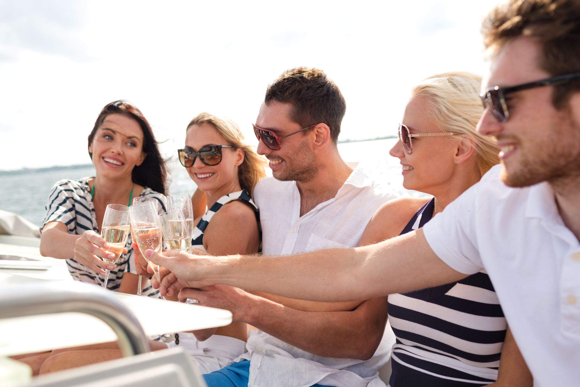 Friends in casual clothing toast with glasses of champagne on a boat deck, smiling and looking toward each other.