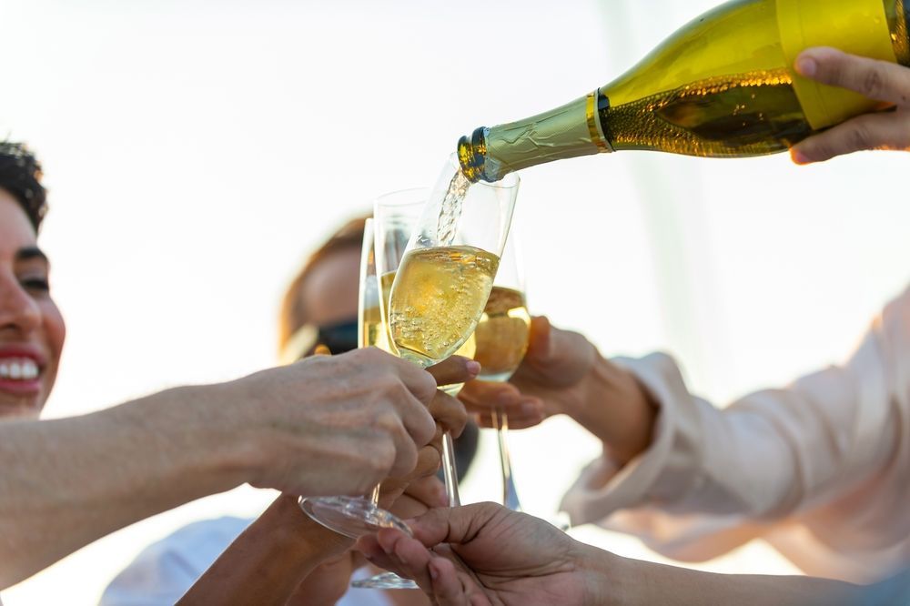 Sparkling wine is poured into several glasses held up by people in a festive, bright outdoor setting.