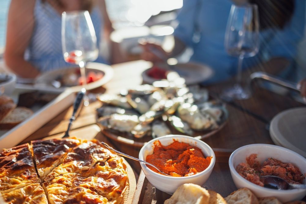 A blurred view of people dining outdoors with a platter of oysters, a crusty tart, and small bowls of dips on a table.
