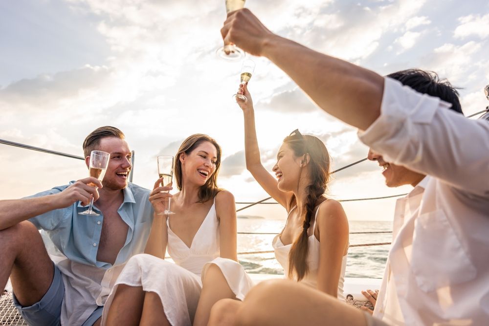Four friends laughing and toasting with champagne glasses on a boat deck during a sunny day.