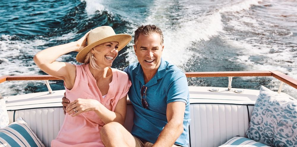 A smiling couple sits together on a boat, traveling across water with a white wake visible behind them.