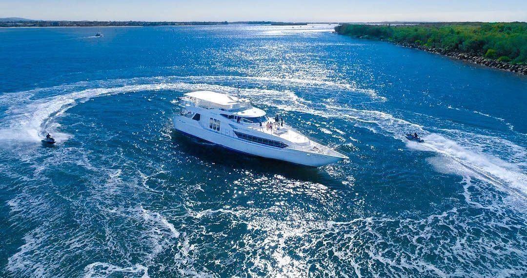 An aerial view of a white yacht surrounded by two jet skis creating circular wakes in bright blue water near a coastline.