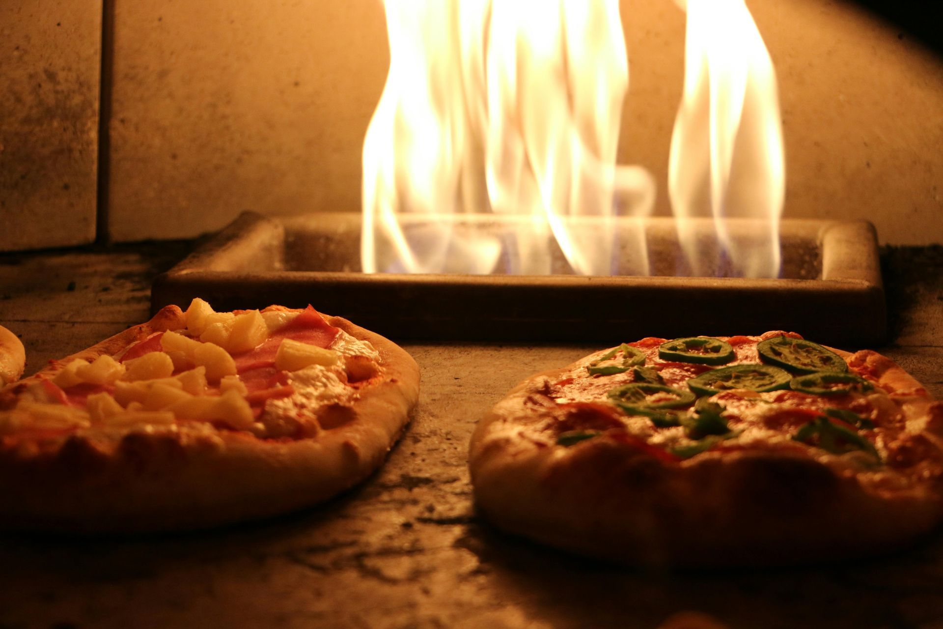 Two pizzas cooking in a wood-fired oven with visible flames in the background.