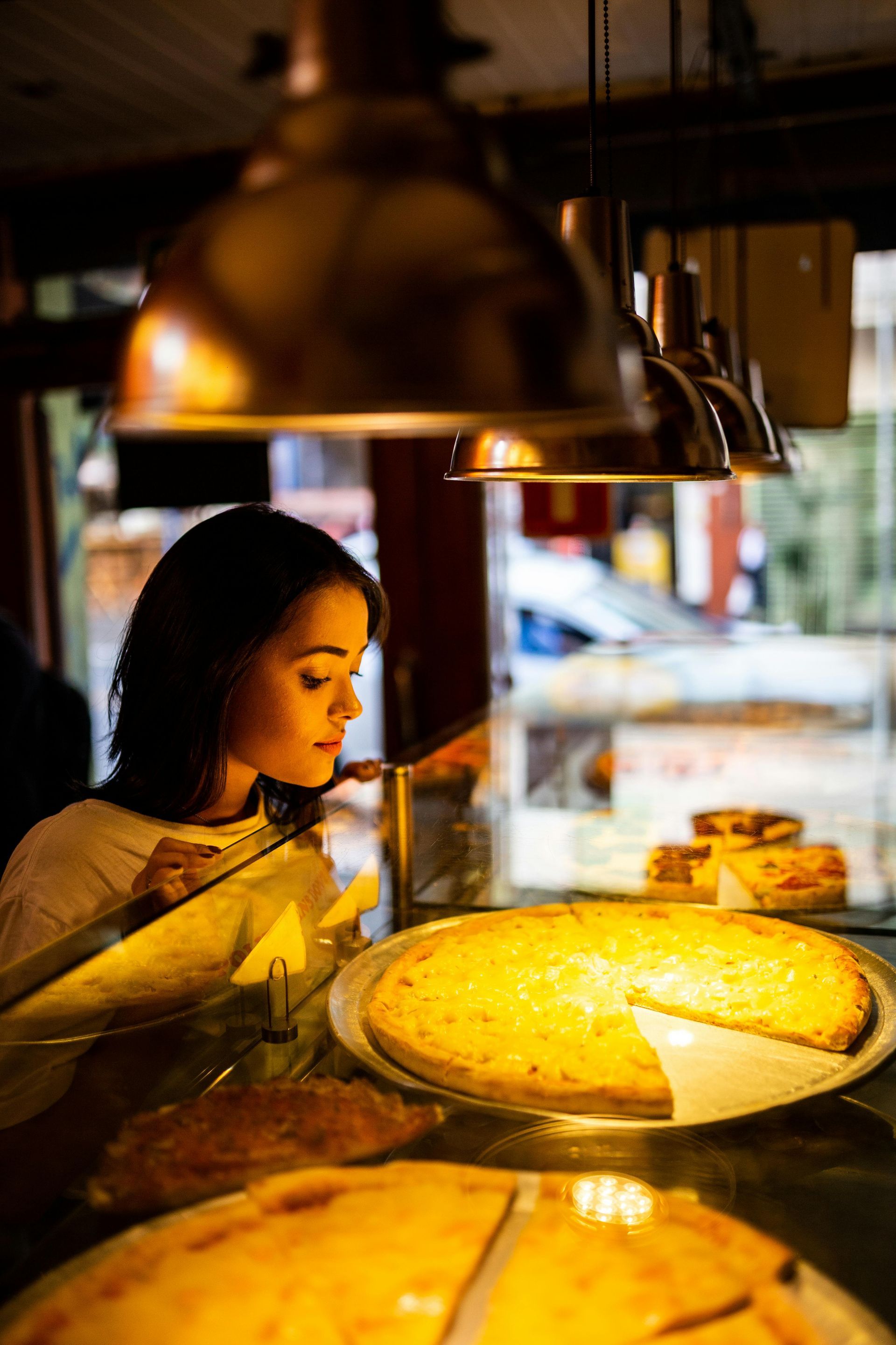Woman looking at pizza on display in a bakery, lit by warm overhead lighting.