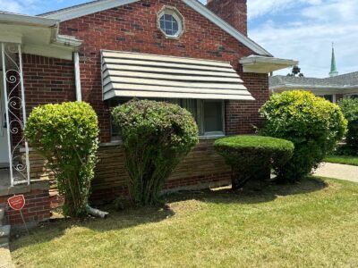 A brick house with a canopy over the window and bushes in front of it.