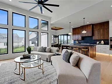 Bright modern open-plan living room with large windows, gray sofas, and a kitchen in the background
