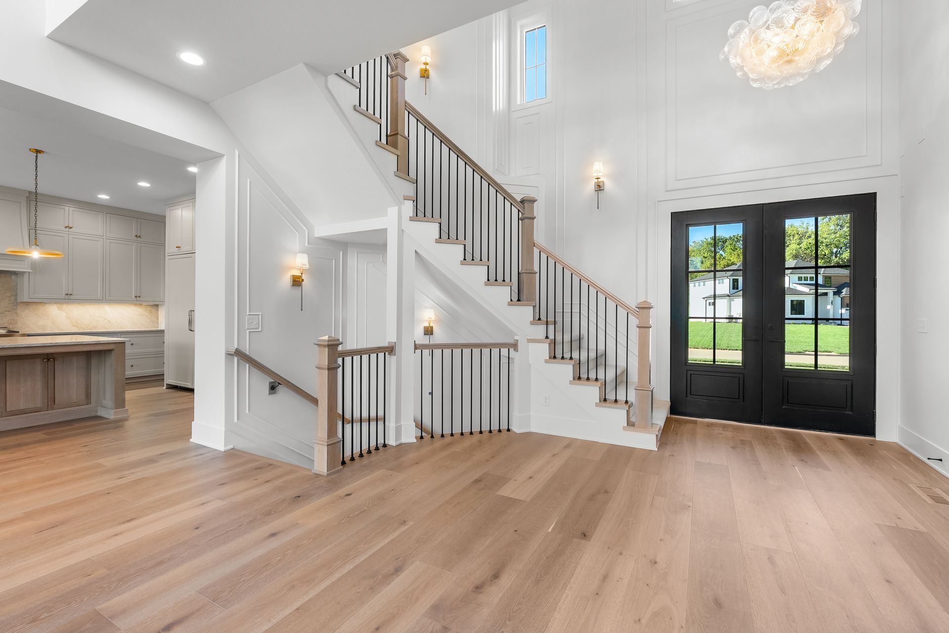 Spacious entry with wood floors, staircase, black double doors, and a glimpse of the kitchen.