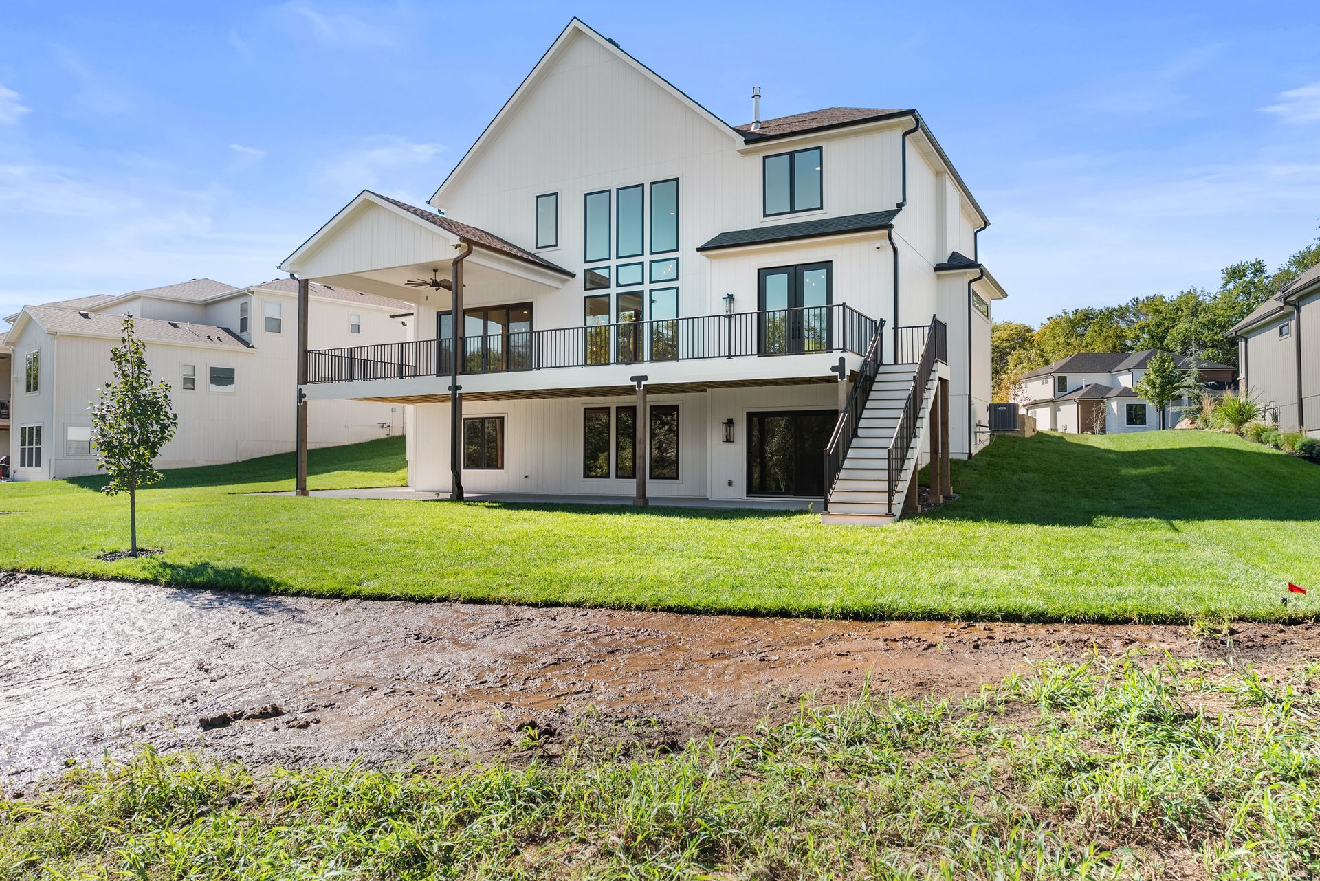 White two-story house with a deck, overlooking a grassy backyard and a pond, under a blue sky.