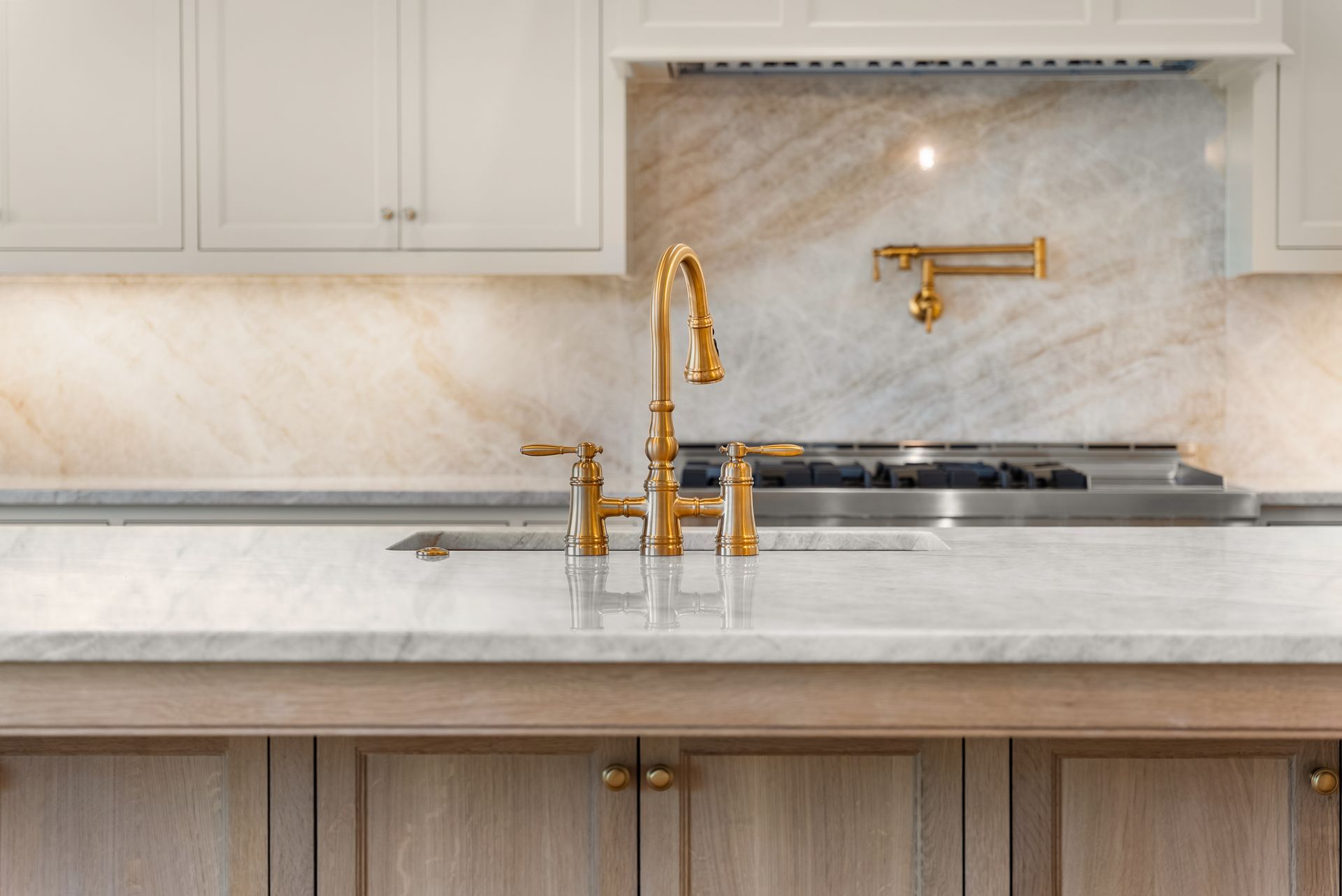 Gold faucet in elegant kitchen with marble backsplash and countertops, light wood cabinets.