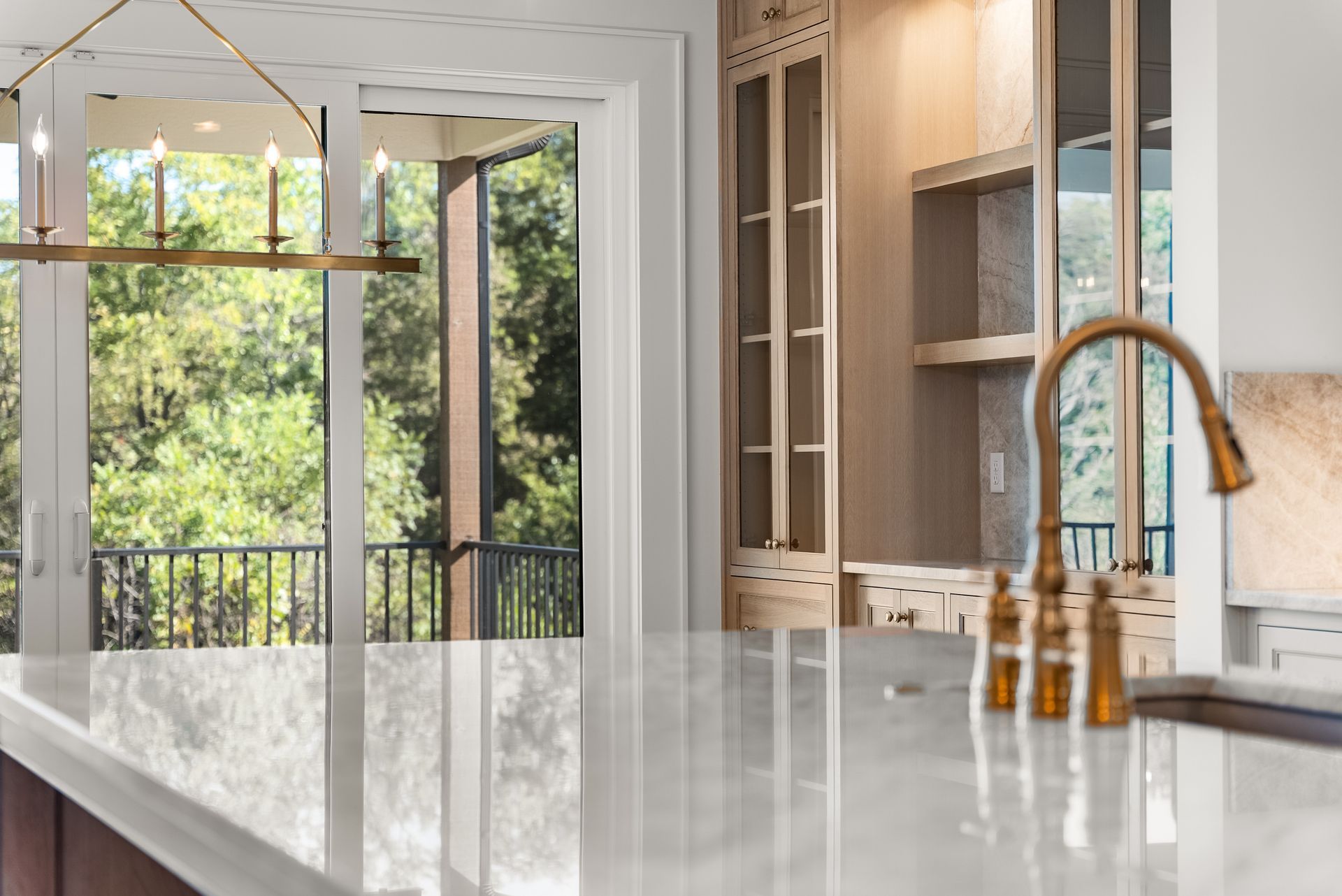 Bright kitchen with gold faucet, marble countertop, and view of trees through a sliding glass door.