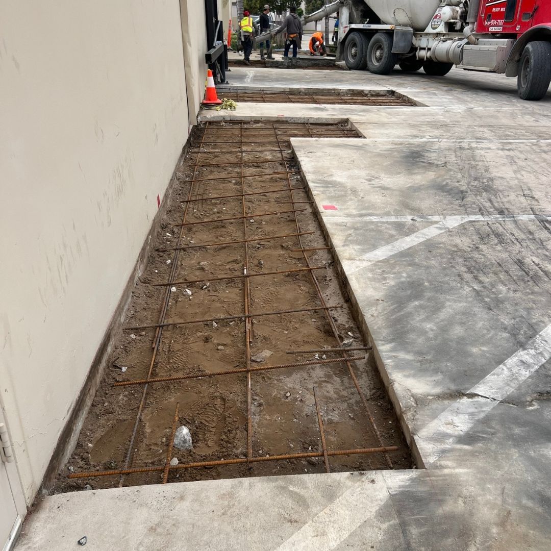 A cement truck is pouring concrete into a parking lot next to a building.