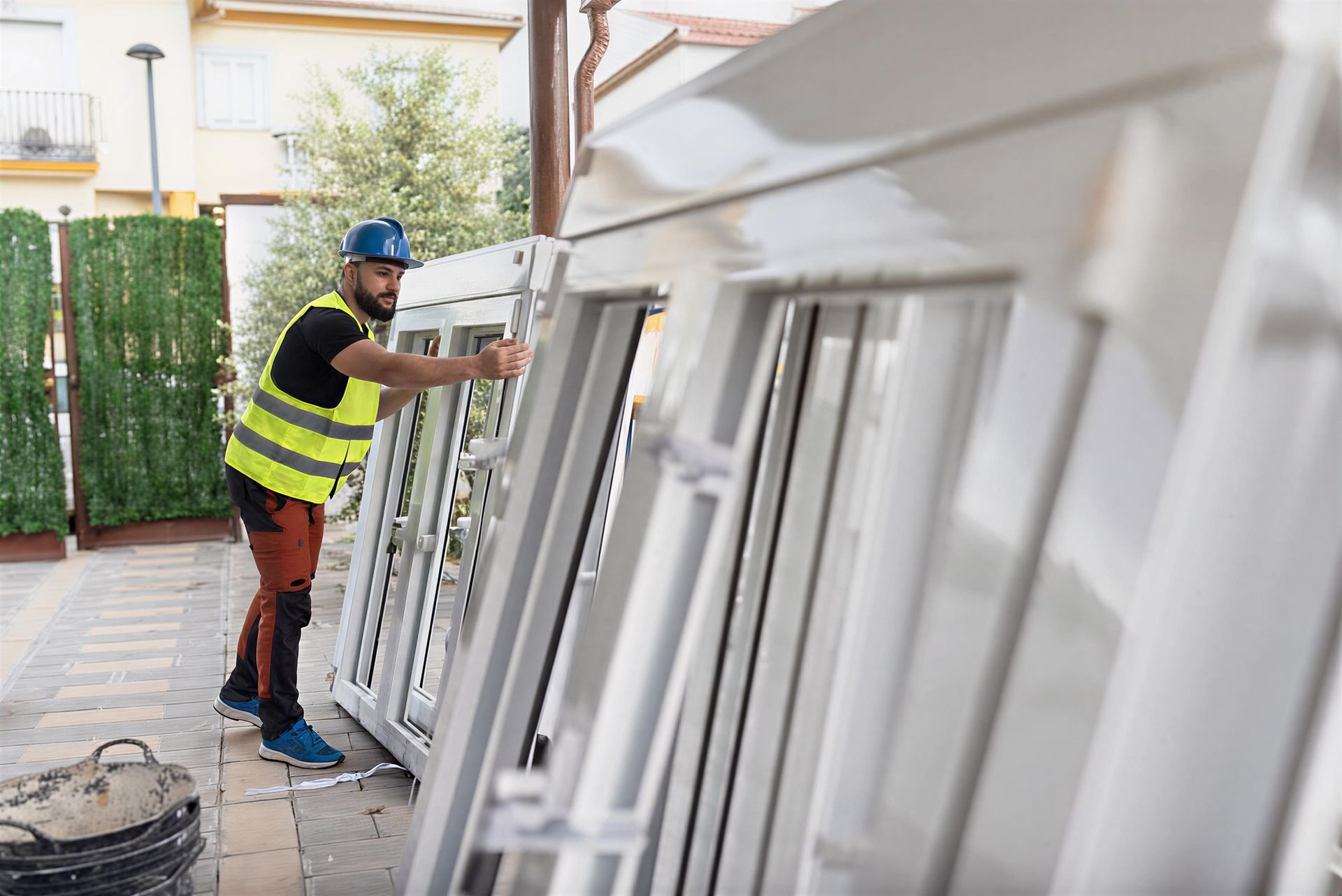 Worker installing large white window frames outdoors on a paved patio area. Worker installing large white window frames outdoors on a paved patio area.