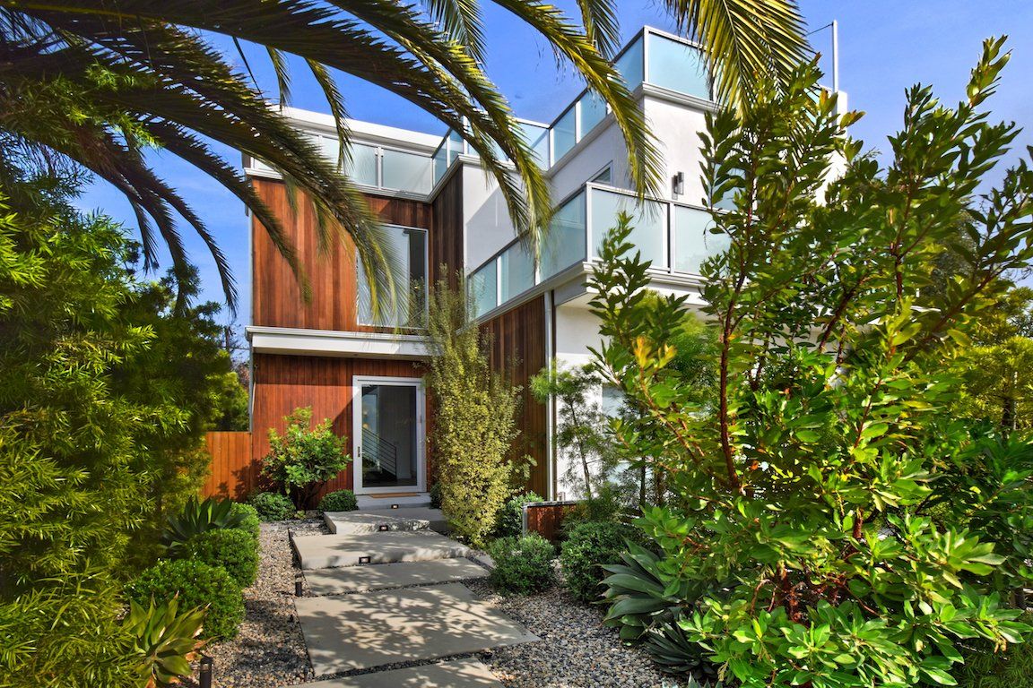 A modern house with a walkway leading to it is surrounded by trees and bushes.