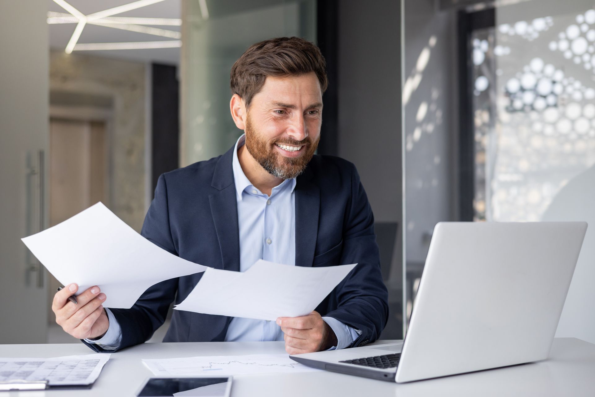 A man is sitting at a desk with a laptop and holding papers.