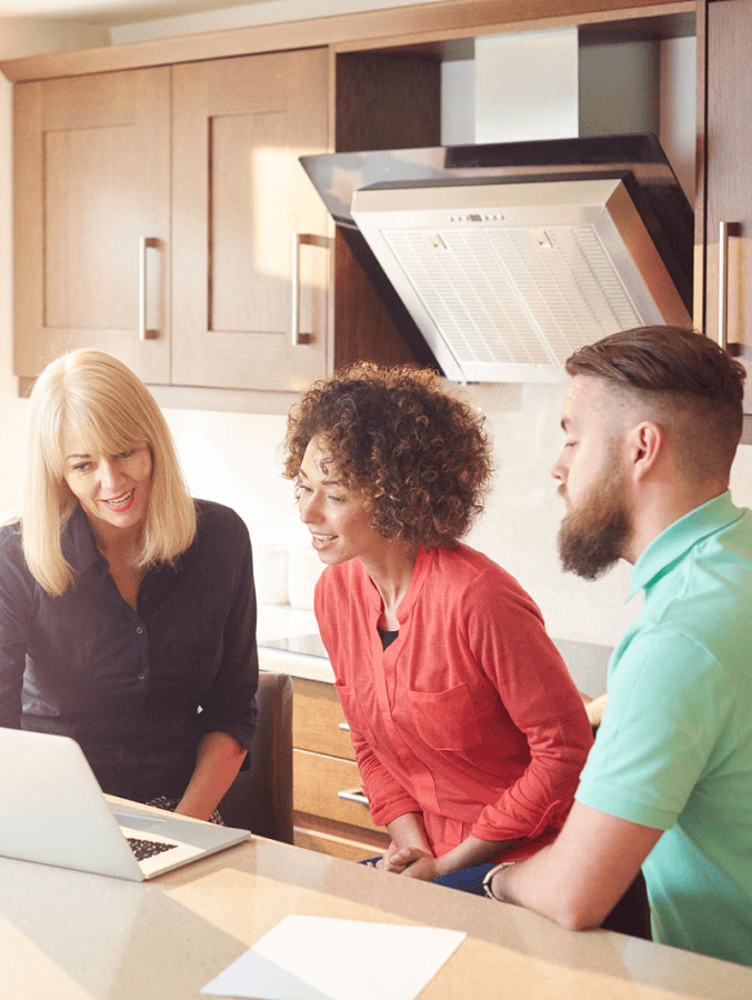 A man and two women are looking at a laptop in a kitchen.