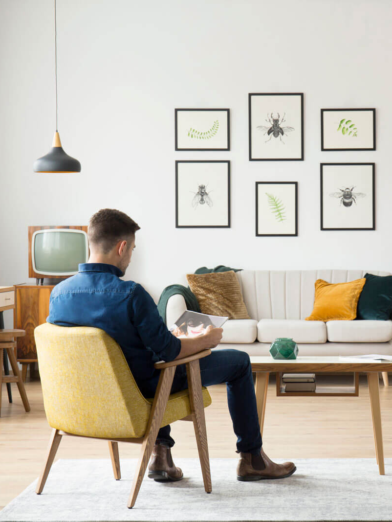 A man is sitting in a yellow chair in a living room reading a book.