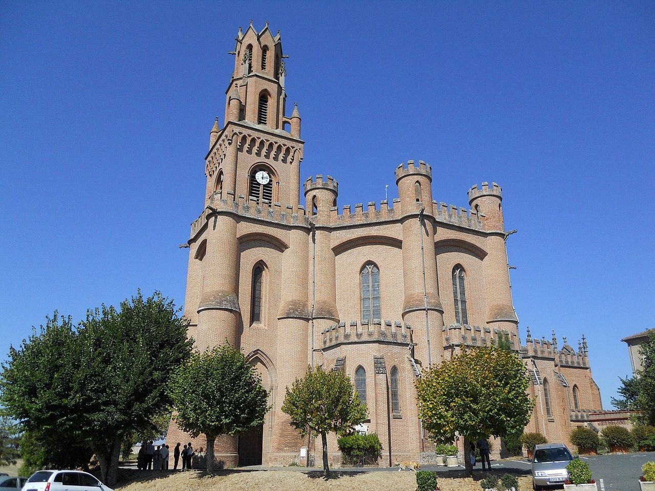 Sous un ciel bleu clair se dresse une église de style gothique en briques beiges, dotée d'un haut clocher et d'un toit crénelé.
