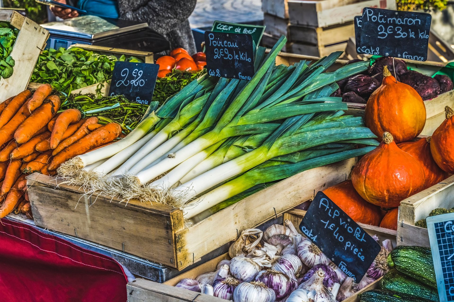 Un étal de marché animé présentant des produits frais, notamment des carottes, des poireaux, des citrouilles et de l'ail, dans des caisses en bois.