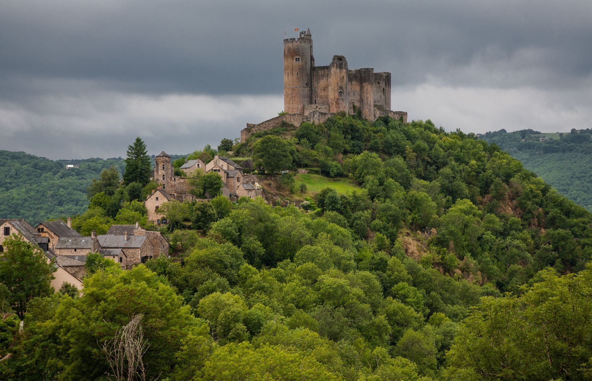 Un château de pierre se dresse au sommet d'une colline verdoyante et arborée, sous un ciel nuageux, dominant un petit village au premier plan.