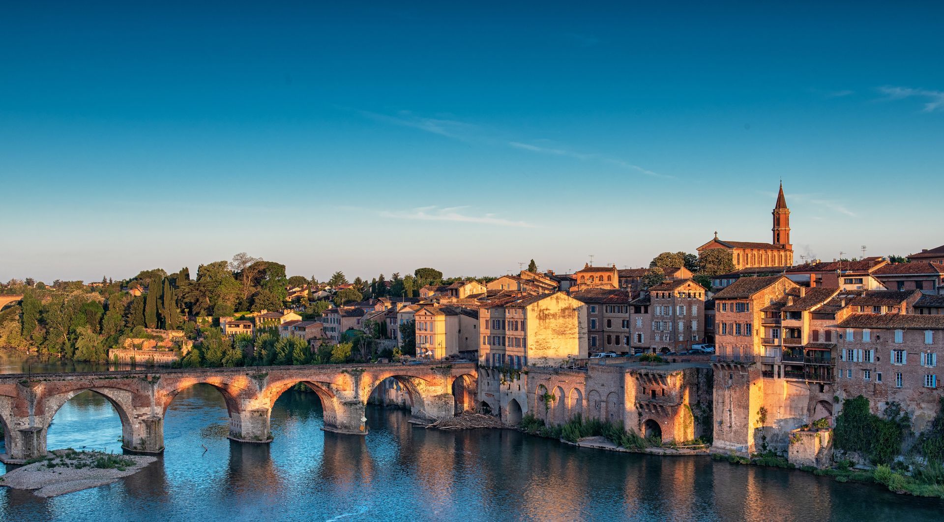 Un vieux pont de pierre enjambe une rivière sous une ville aux toits de tuiles et au clocher, au coucher du soleil.