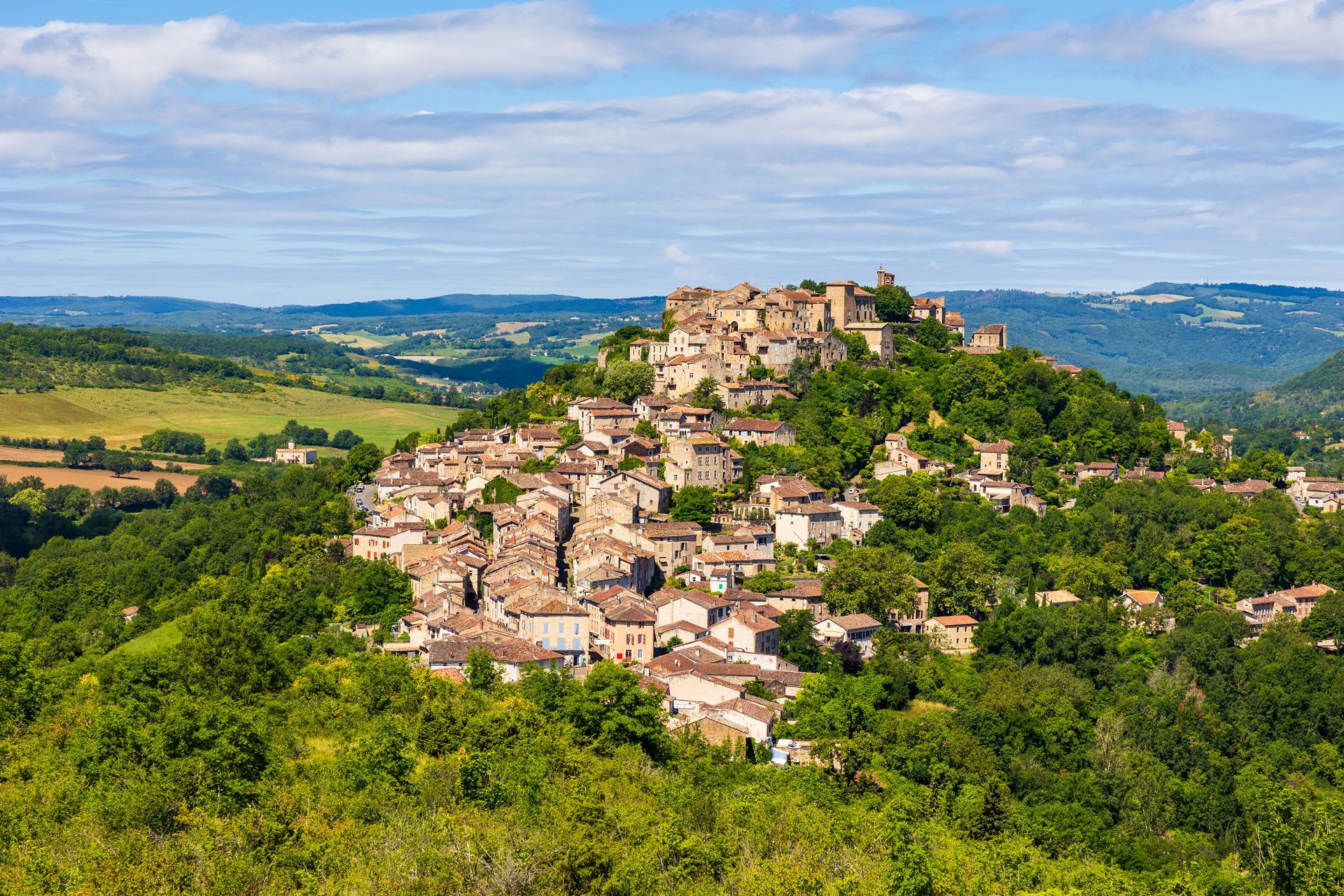 Un village de pierre aux toits de tuiles rouges, perché sur une colline boisée et luxuriante, sous un ciel d'un bleu éclatant parsemé de nuages épars.