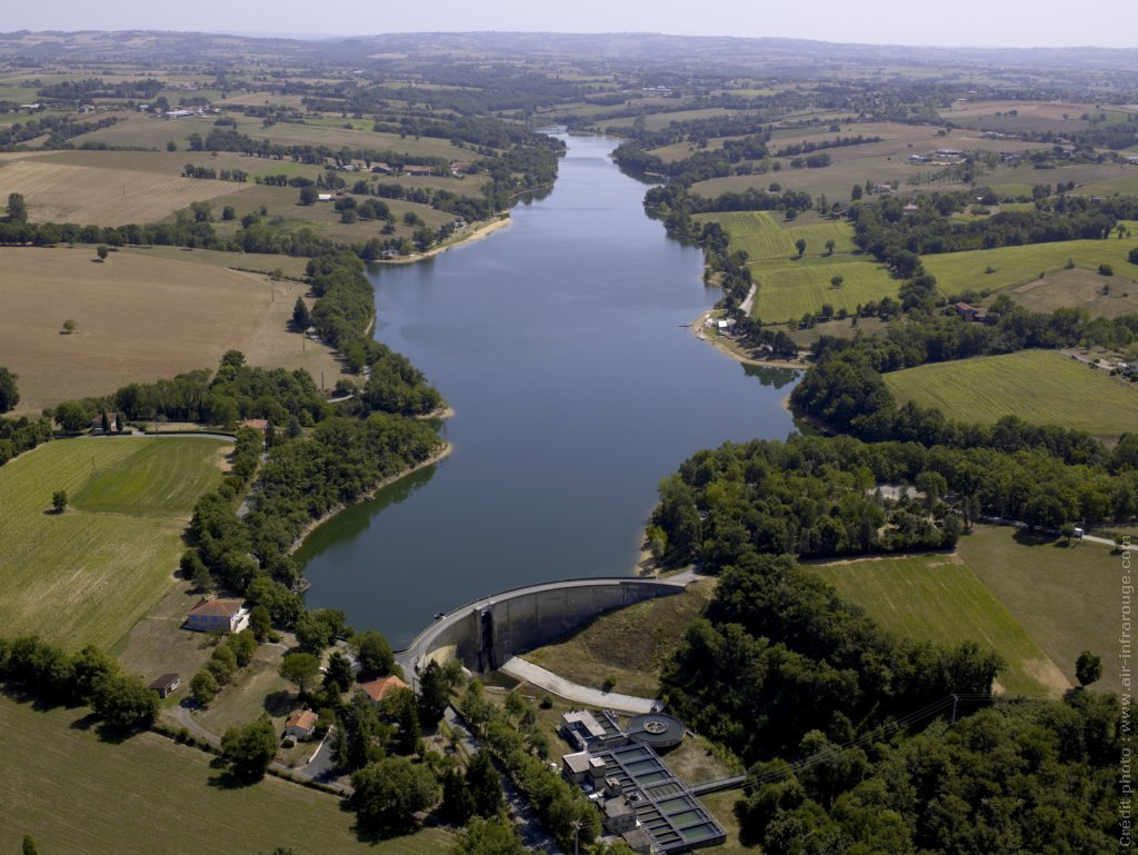 Vue aérienne d'un barrage incurvé retenant un grand réservoir calme, entouré de champs verdoyants et de forêts luxuriantes.