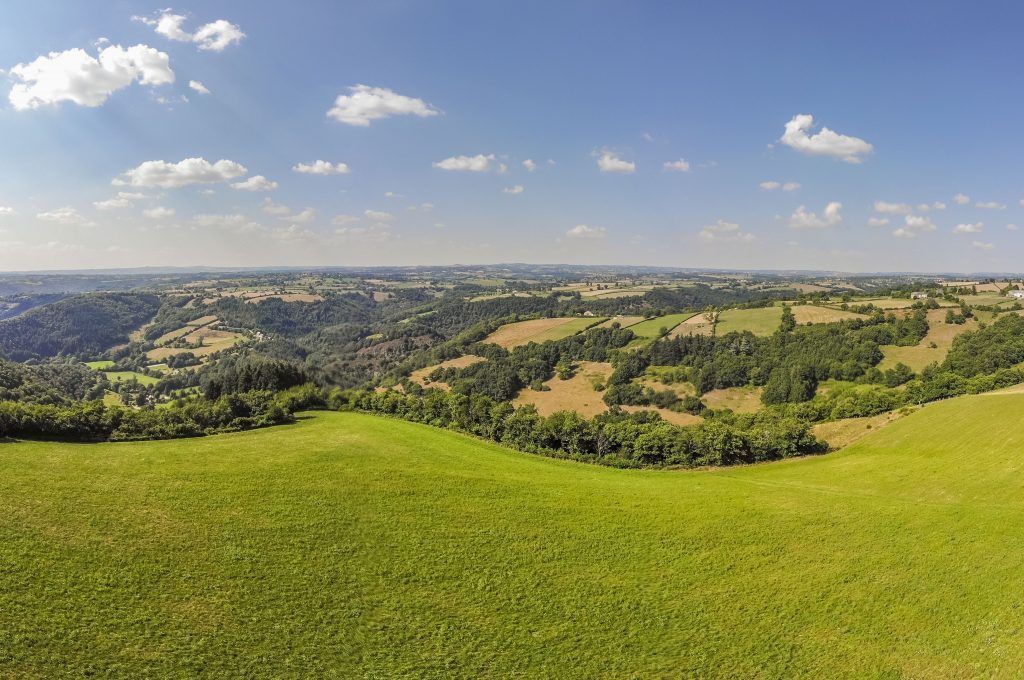 Vue panoramique sur des collines et des champs verdoyants sous un ciel bleu éclatant parsemé de quelques nuages légers.