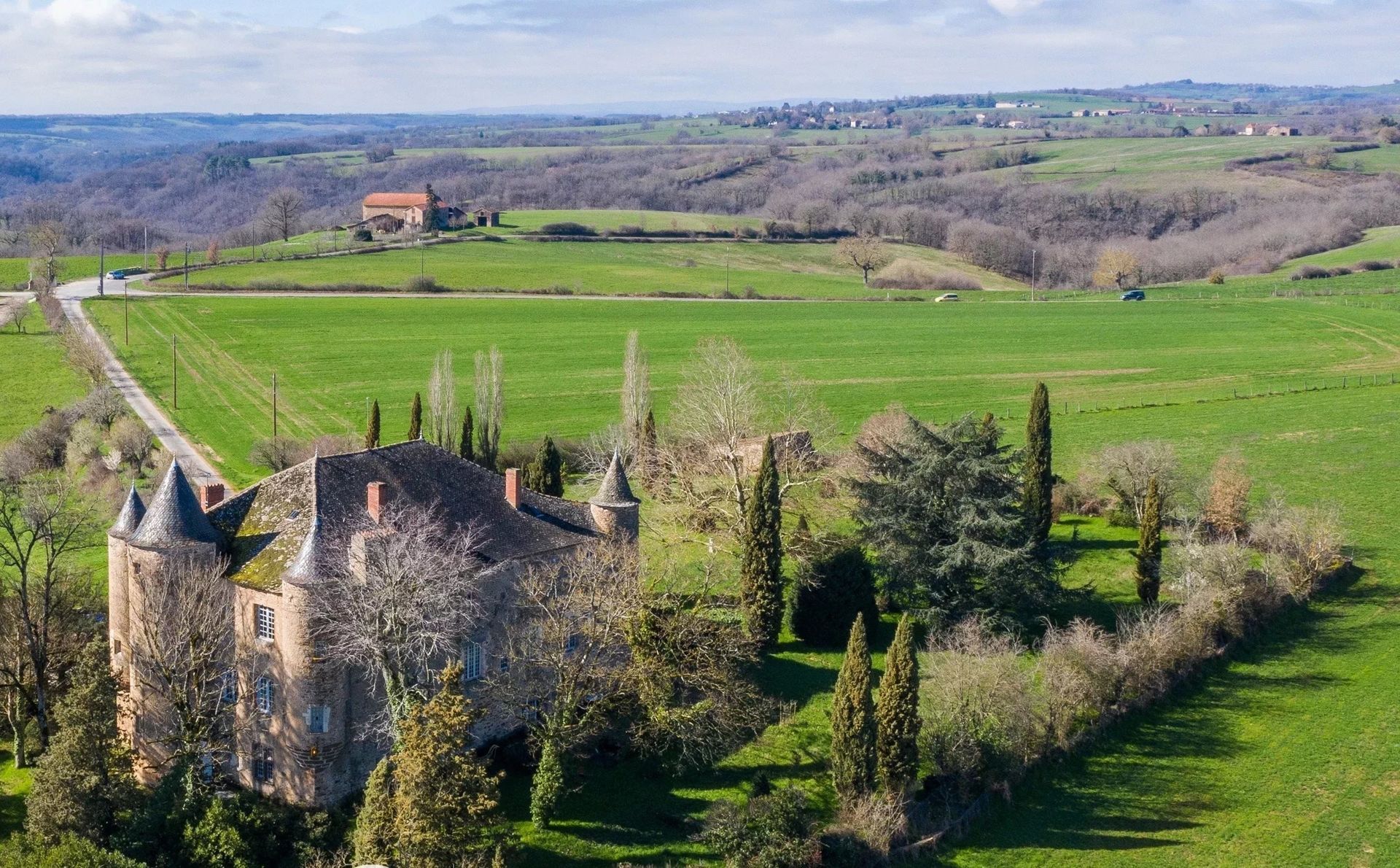 Vue aérienne d'un château en pierre aux tours coniques, entouré de champs verdoyants et d'arbres sous un ciel lumineux.