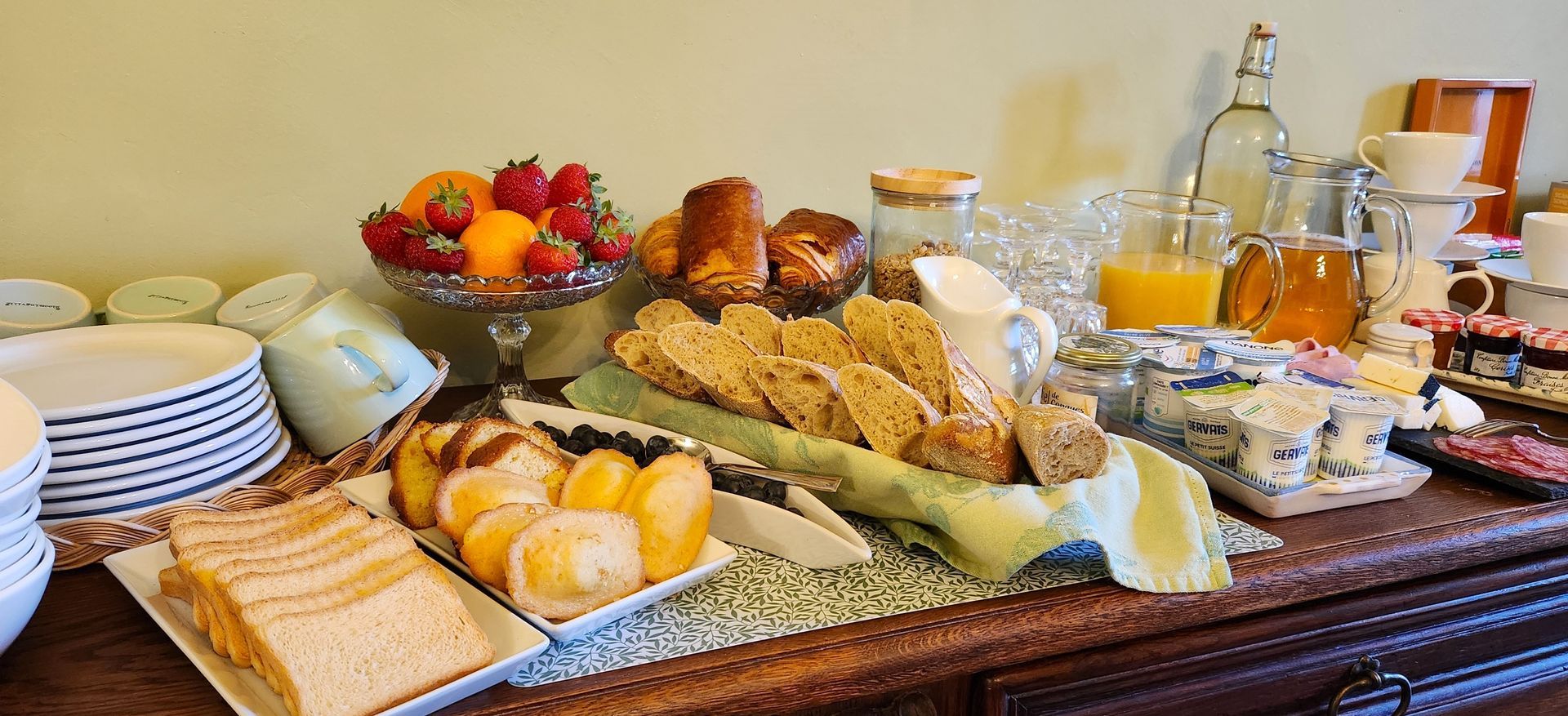 Un buffet de petit-déjeuner composé de tranches de pain, de petits pains, d'une corbeille de fruits, de jus de fruits et de condiments, présenté sur une table en bois.