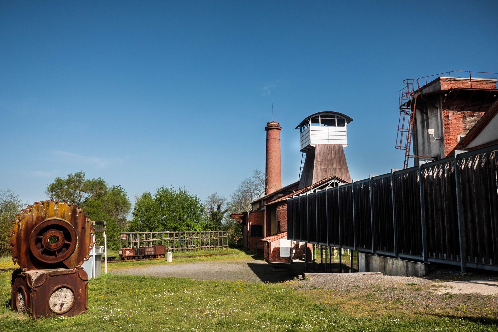 Une ancienne installation minière rouillée, avec une haute cheminée en briques, une tour de guet blanche et une grande roue métallique circulaire rouillée.
