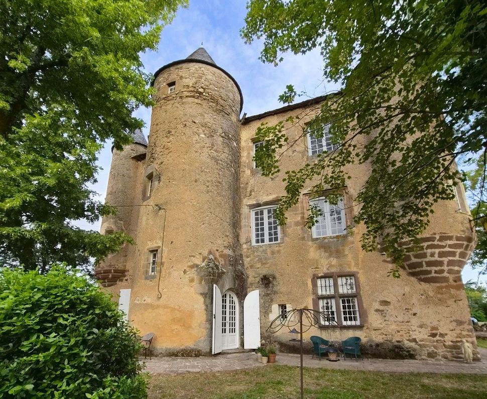 Un vieux château en pierre avec deux tours rondes, des portes blanches et des fenêtres cintrées, entouré d'arbres et d'une cour gazonnée.