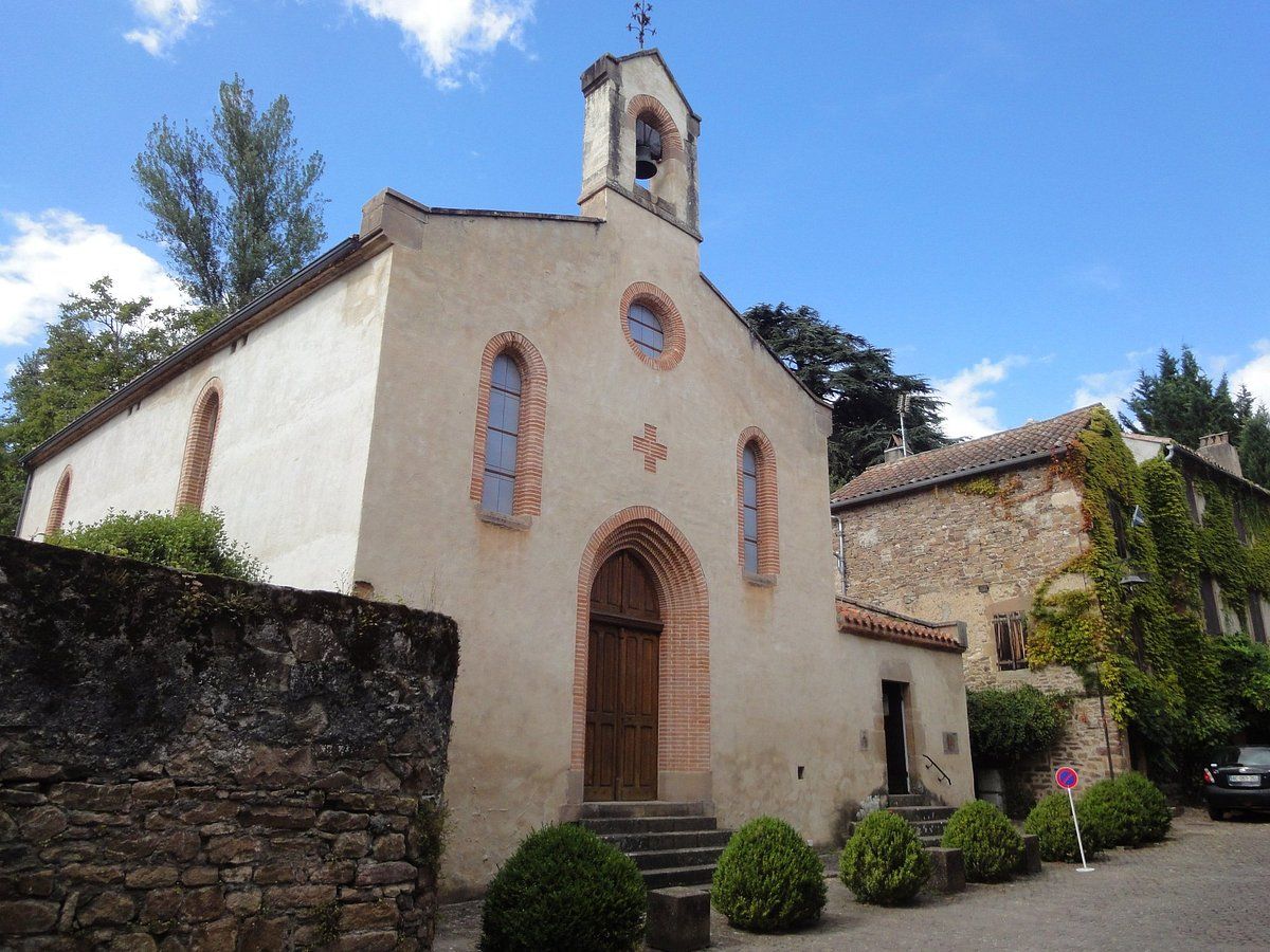 Une église en pierre avec un clocher et une entrée voûtée, bordée de petites haies, sous un ciel bleu parsemé de nuages blancs.